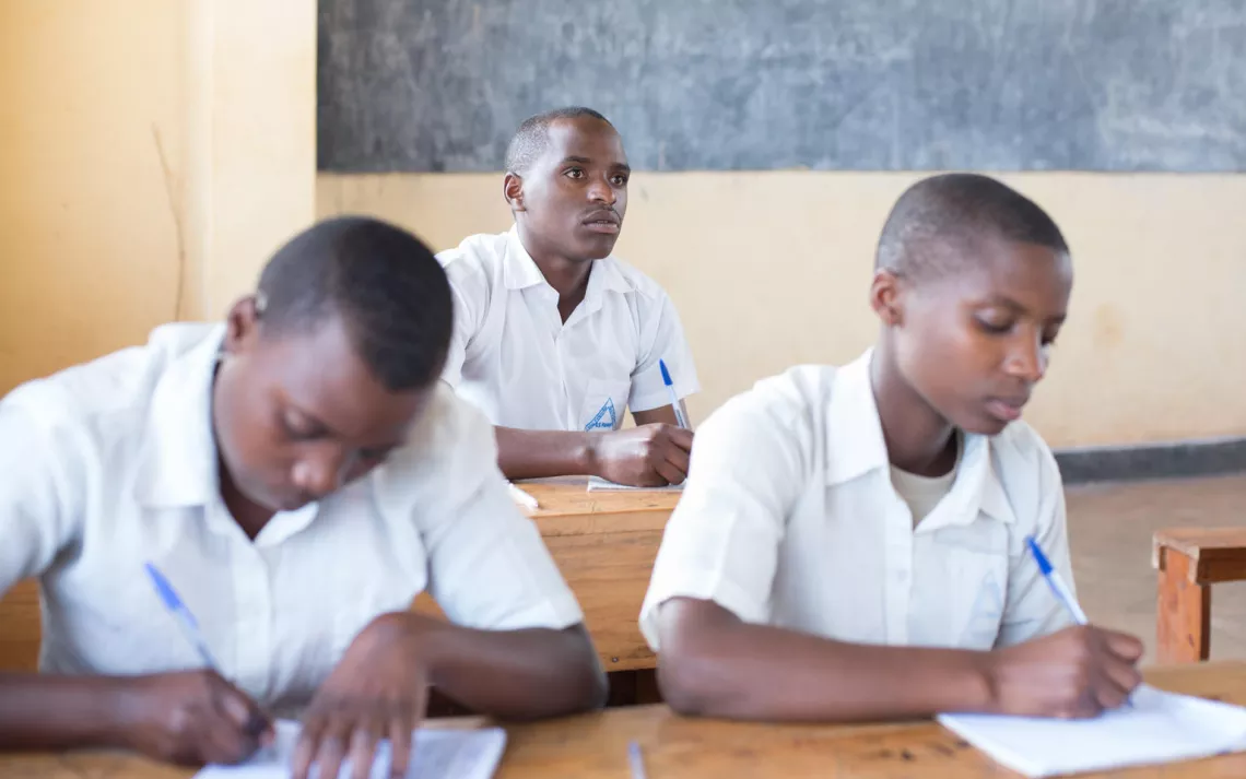 Boy student in Rwanda classroom writes in book and looks at teacher
