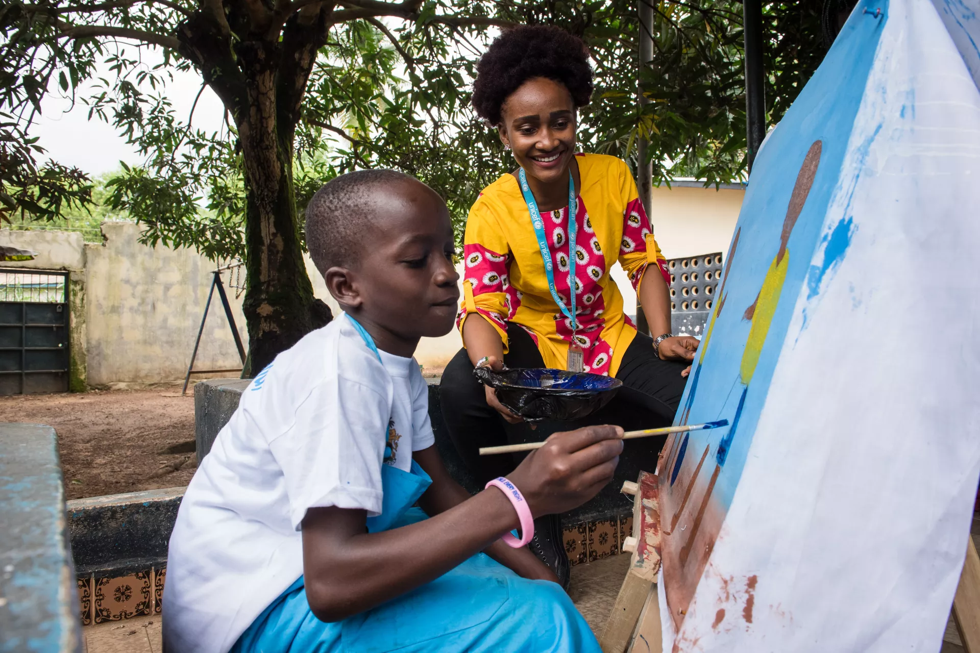 A child paints a drawing during an art session in Bo, southern Sierra Leone as UNICEF Communication Officer looks on.