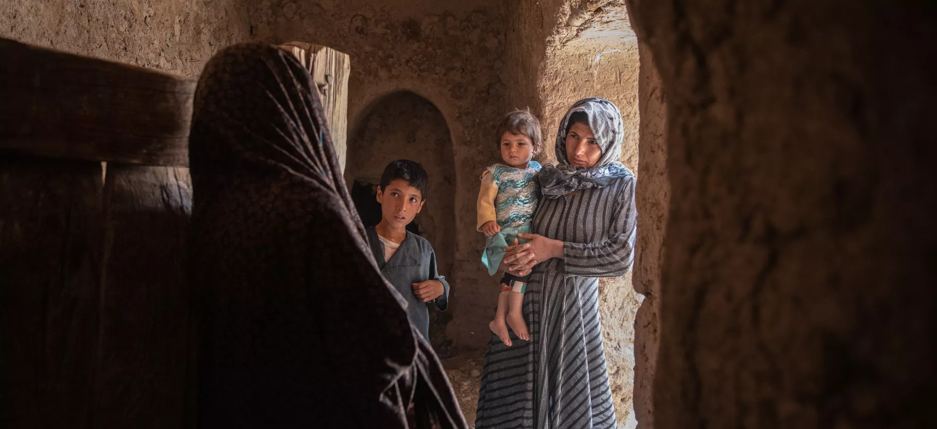 A mother carrying a small child and holding the hand of her young son beside her, stand in the doorway of a home, facing a volunteer vaccinator.