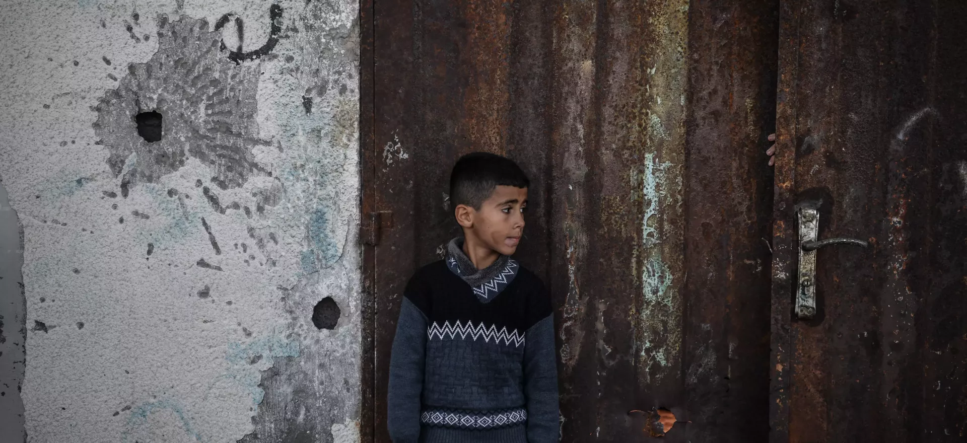 Un ni&ntilde;o se encuentra junto a una pared acribillada a balazos y una puerta oxidada en Gaza