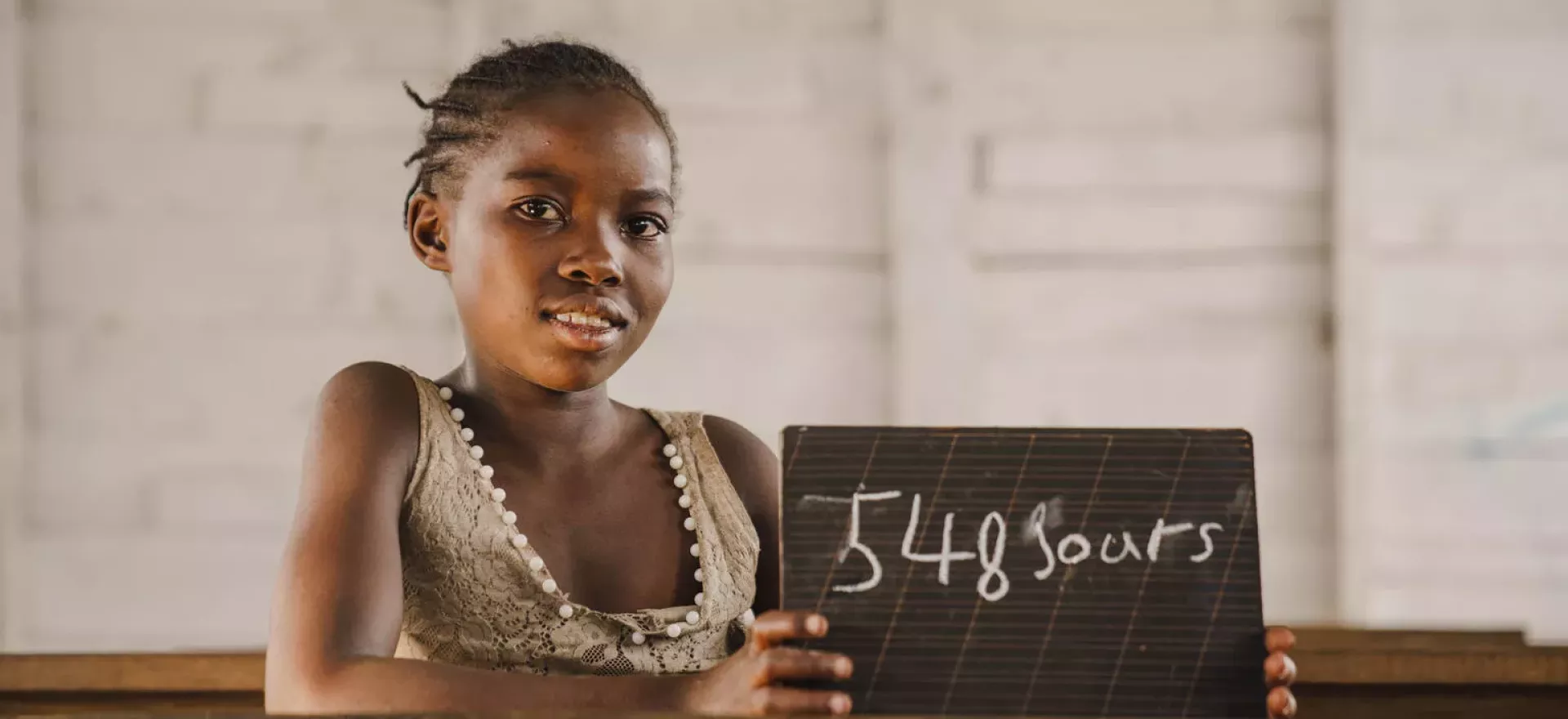 Democratic Republic of the Congo. A girl sits at a desk holding a chalkboard showing how many days she has been out of school.