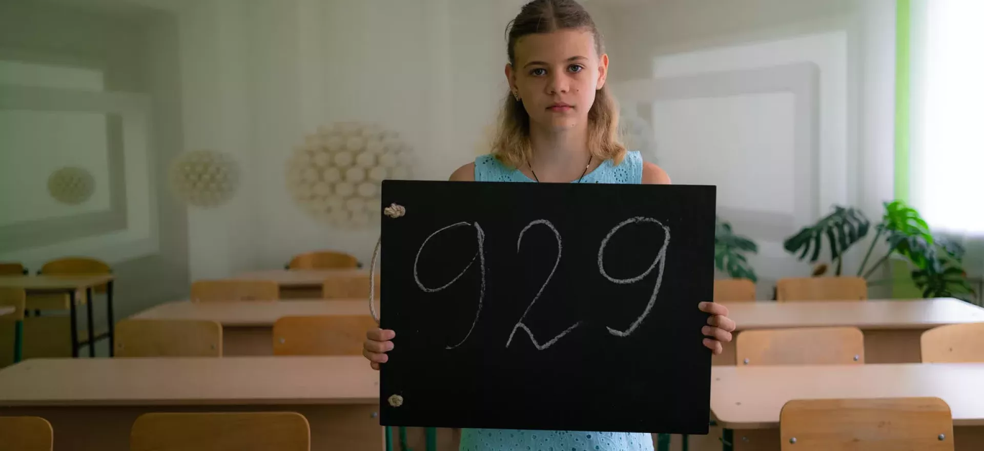 Ukraine. A girl stands in an empty classroom holding a chalkboard showing the number of days she has been out of school.