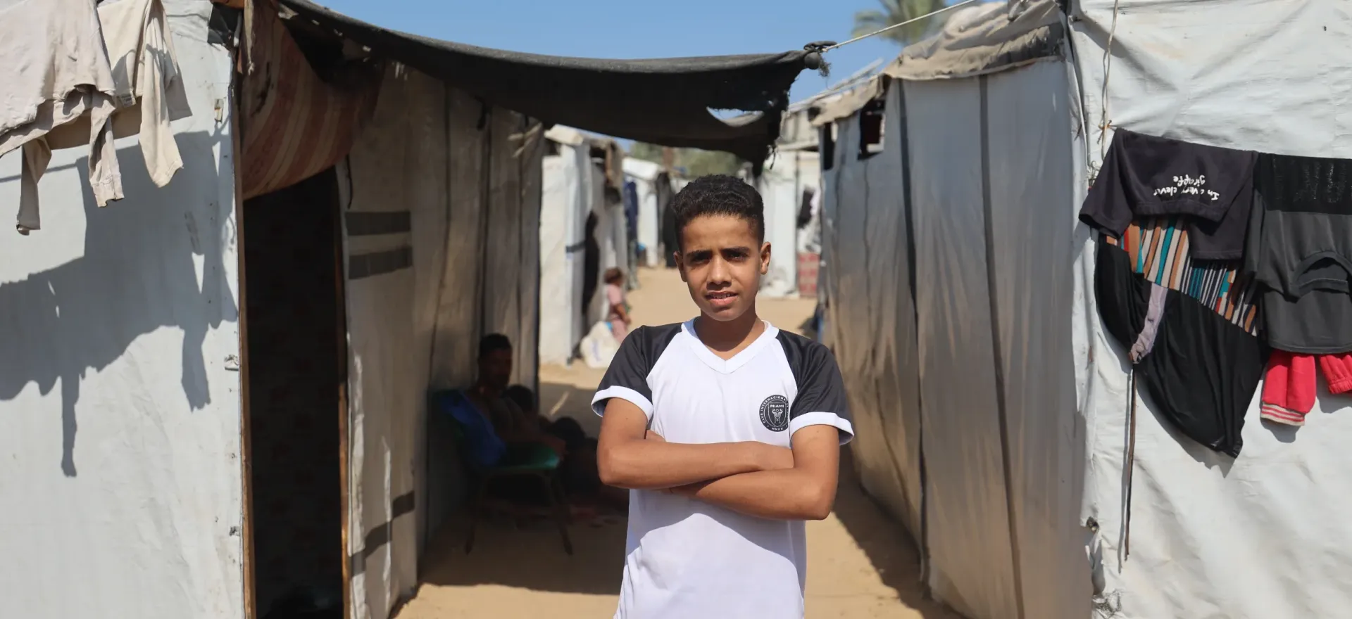 Gaza. A boy stands with his arms folded outside some tents.