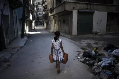 Syria. A girl, carrying jerrycans of water, walks past a pile of debris.