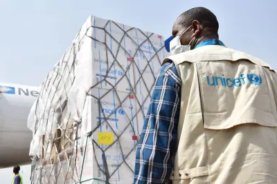 A man wearing a mask and a UNICEF jacket standing in front of a shipment of emergencies supplies being loaded into a plane