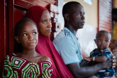 A young girl sits next to her mother with her little brother sitting on her father's knee next to her mother.