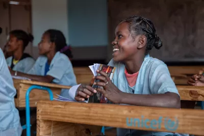 Students in a classroom, seated at wooden desks with "unicef" written on them. One student in focus smiles while holding notebooks.