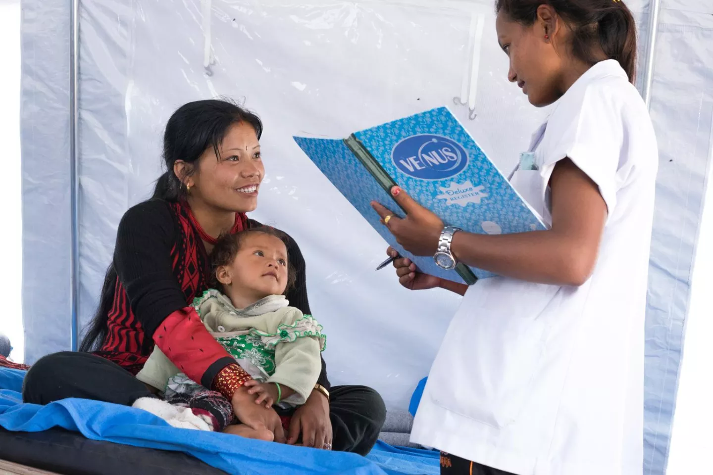 Sangeeta Thami, sitting on a bed with her 1-year-old daughter, Sujata, in her lap, chats with staff nurse Swosthani Shrestha