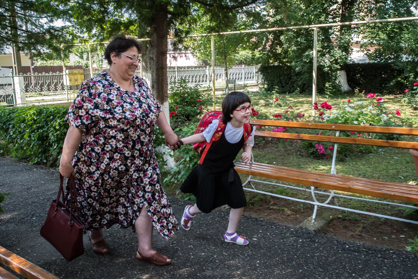 A young girl with a backpack runs to school with a smile, eagerly dragging her guardian behind.