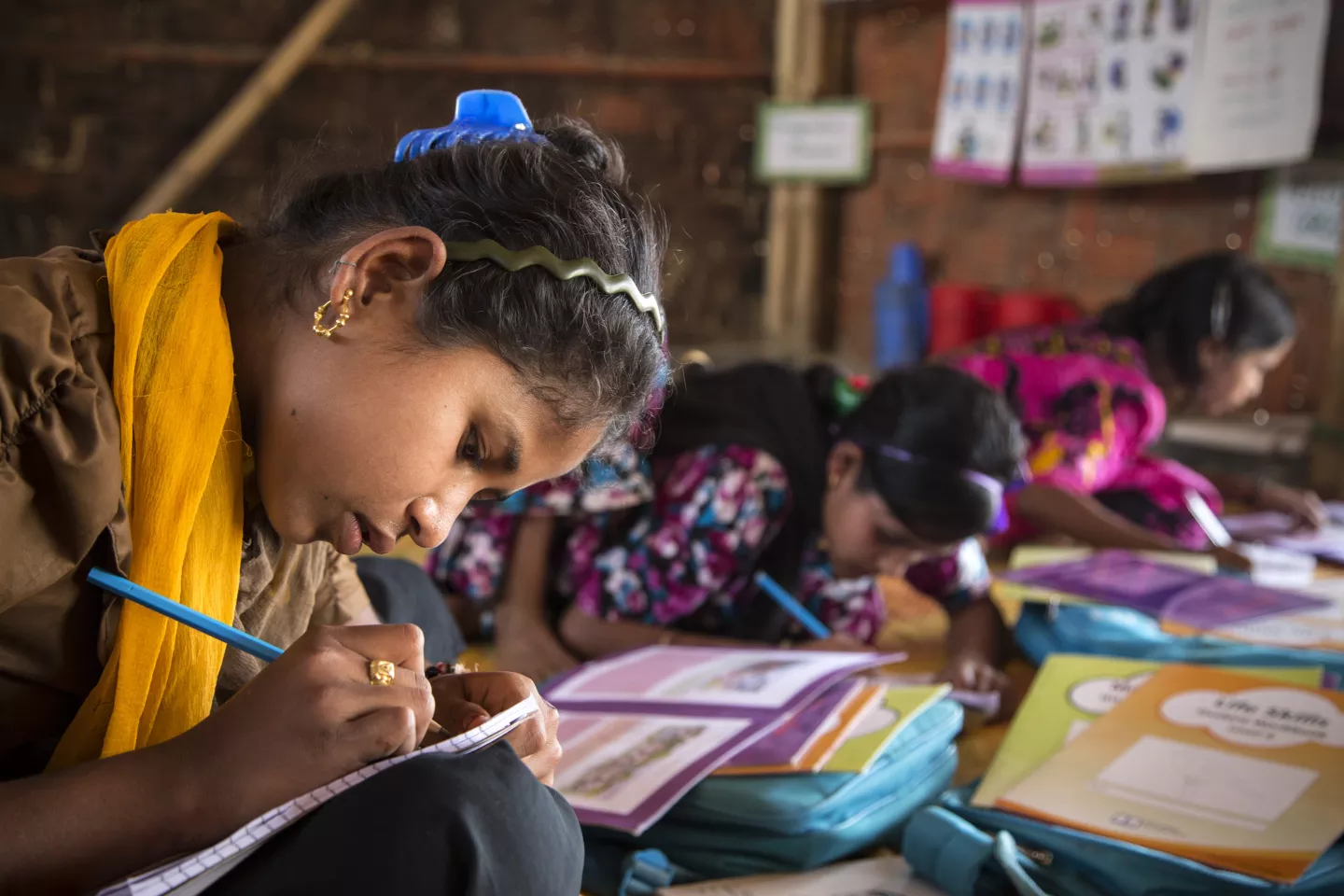 A girl stoops over her lap, writing diligently in a pad of paper, and surrounded by colourful books.
