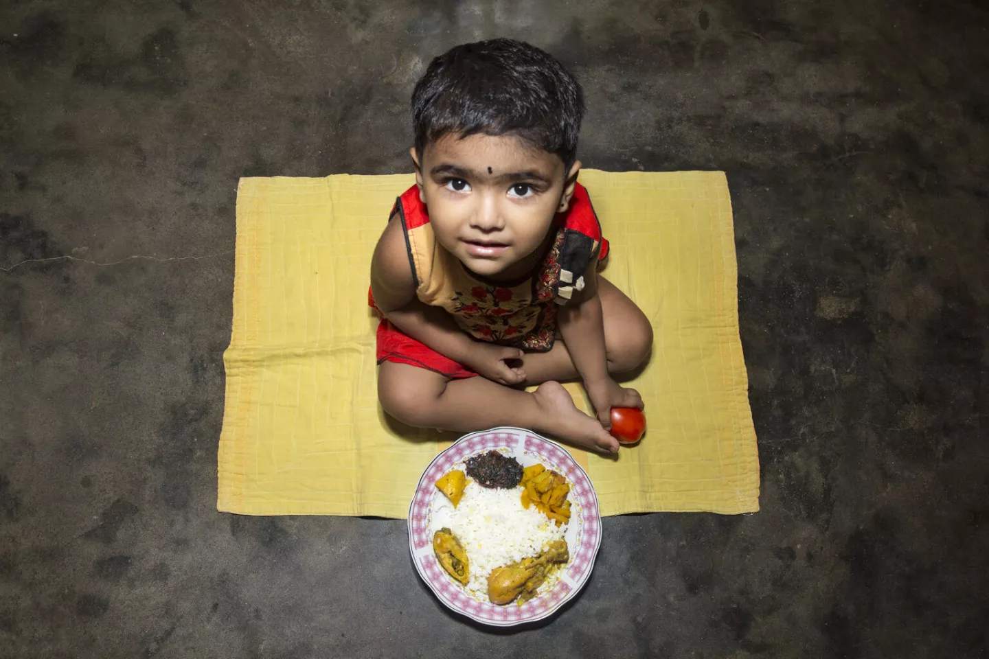 Bangladesh. A boy sits on a rug on the floor next to a plate of food.