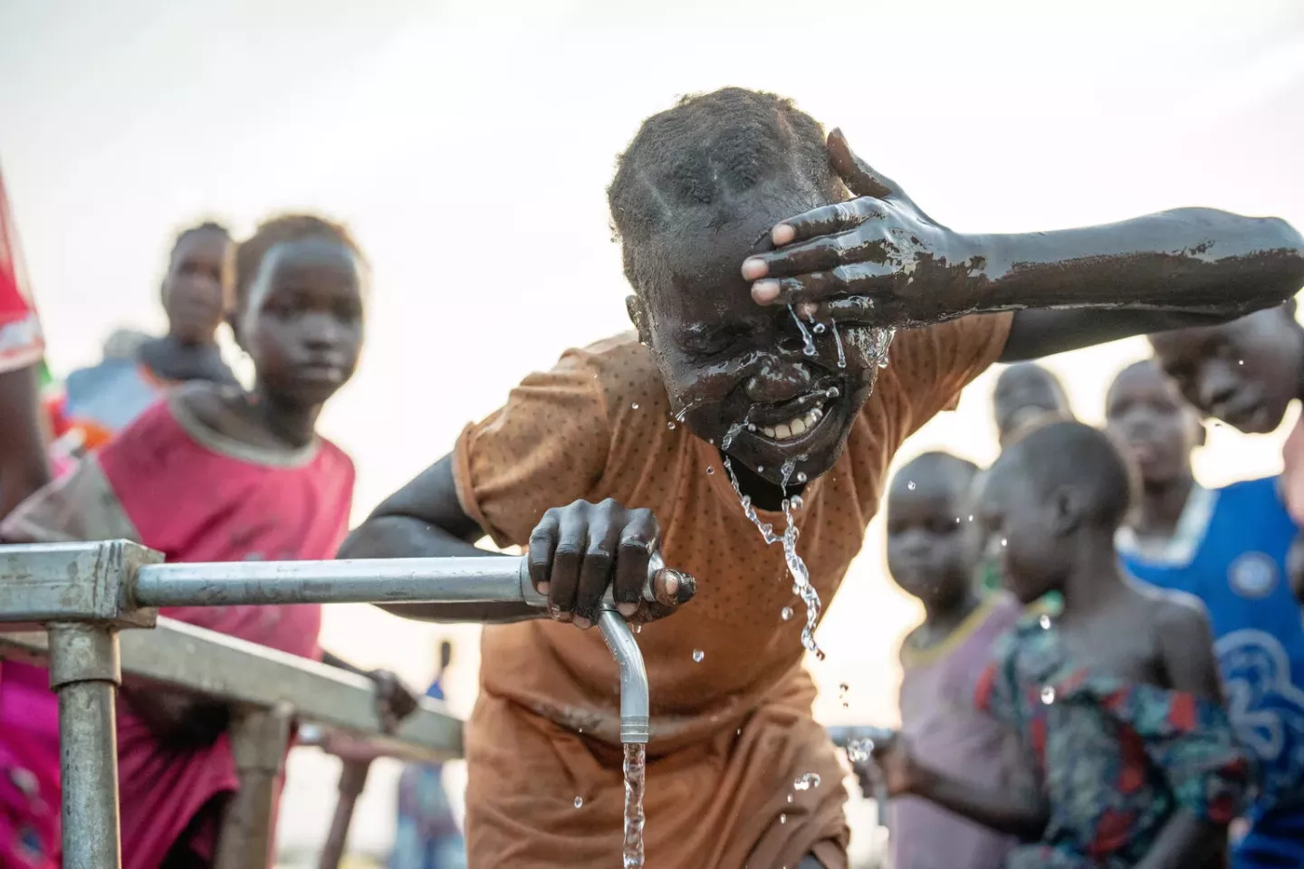A smiling girl splashes water on her face from an outdoor tap.