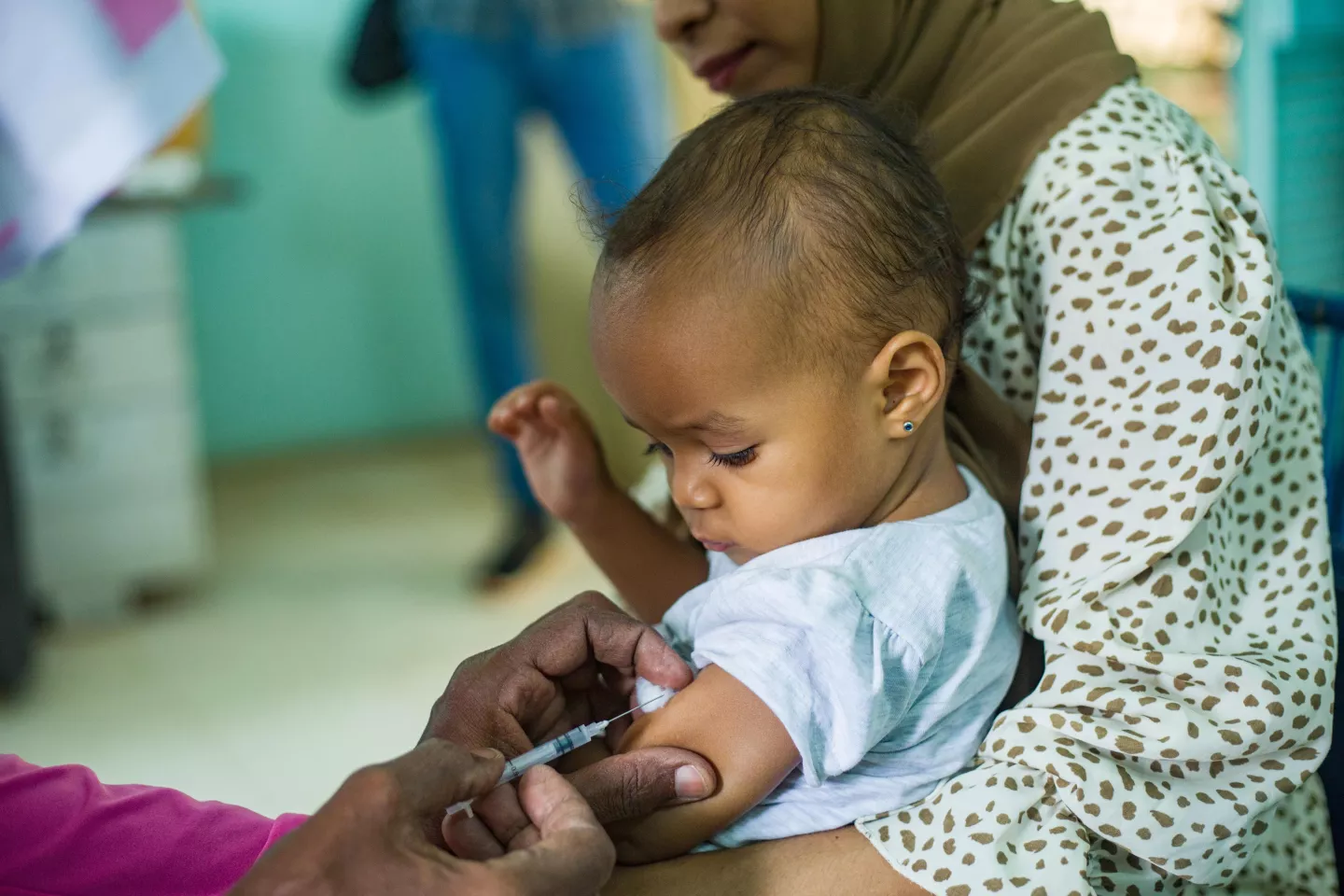 A baby boy sitting on his mother's lap, looks down at his arm where he is about to get vaccinated