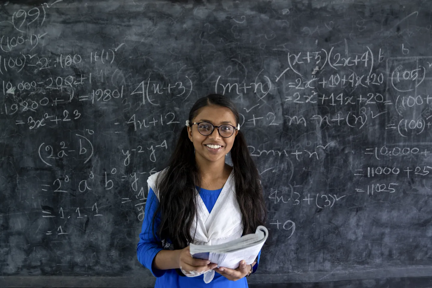 Girl from Bangladesh standing in front of a blackboard with her math books.