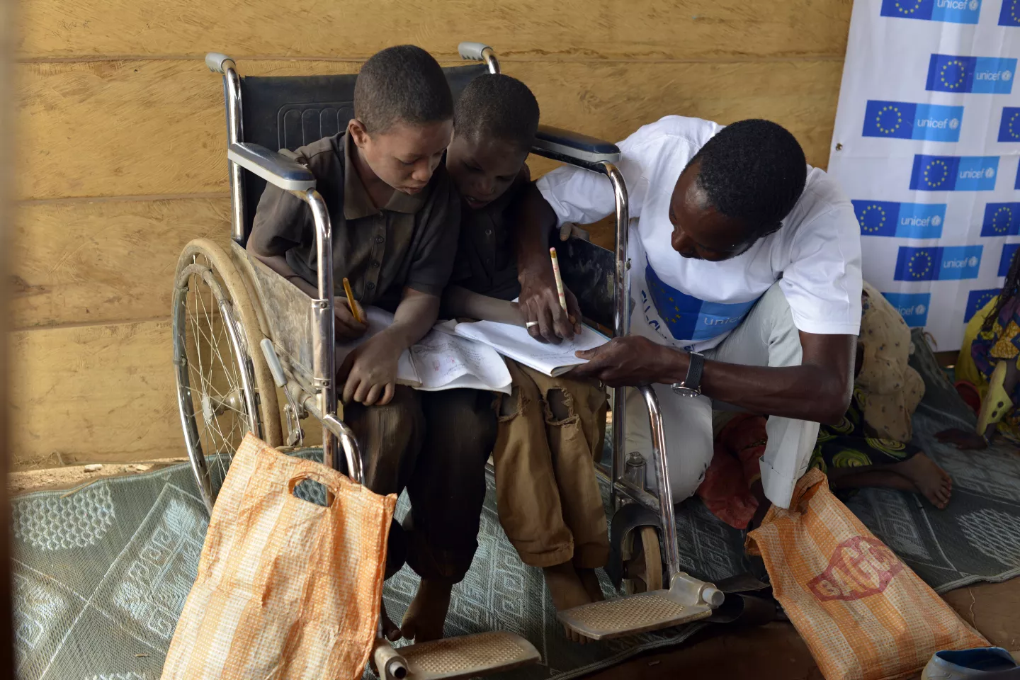 Two boys with disabilities, who are sharing a wheelchair, get help from their teacher during a primary-school class in a learning and protection space at the Gado site for refugees from the Central African Republic, in East Region.