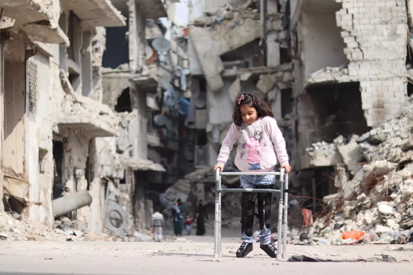 Eight-year-old Rama uses a walker to traverse her neighborhood in east Aleppo, Syria, where the scars of war mark every building behind her. . 