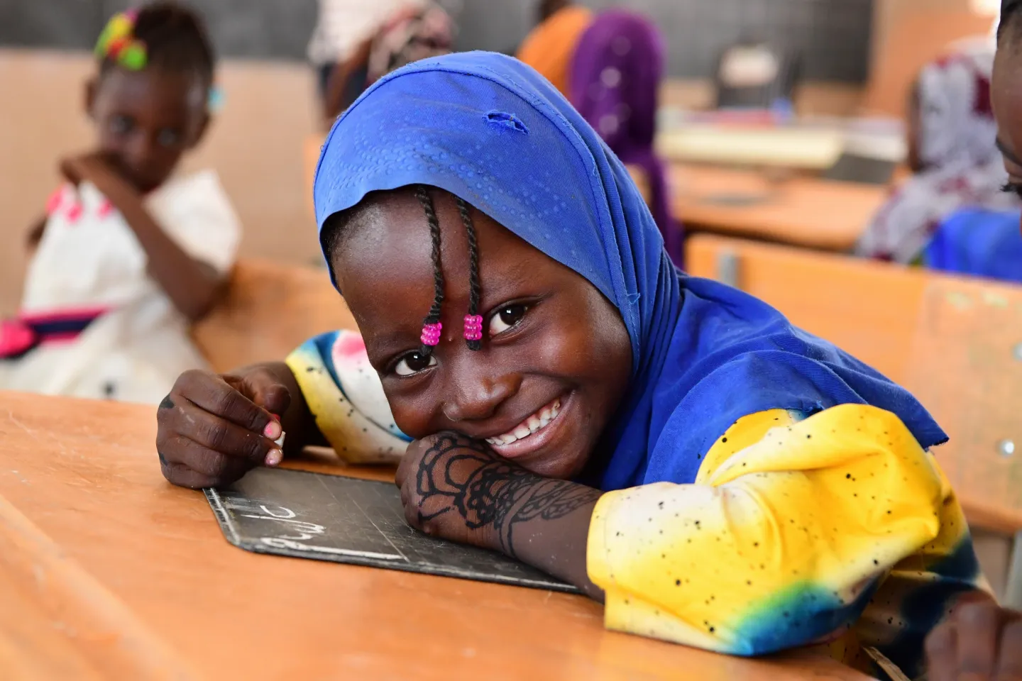 A young girl wearing a blue headscarf, with small plaits with a pink bead on each hanging over her forehead, smiles broadly as she looks up from writing with a piece of chalk on a board on her desk in class.