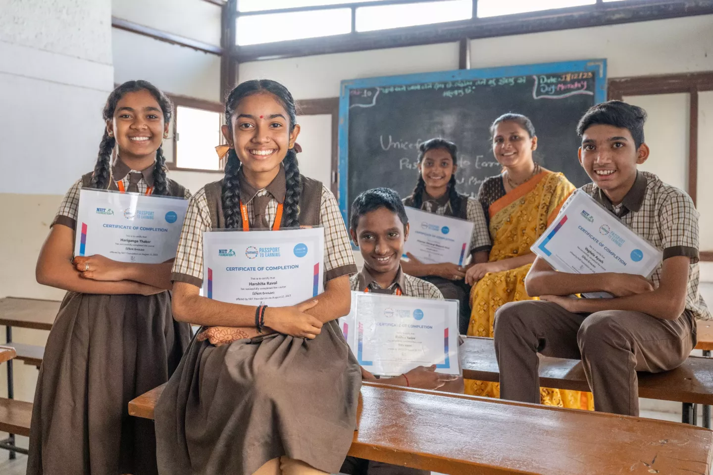 Teen girls and boys dressed in school uniforms sit and stand around a classroom showing off their certificates 