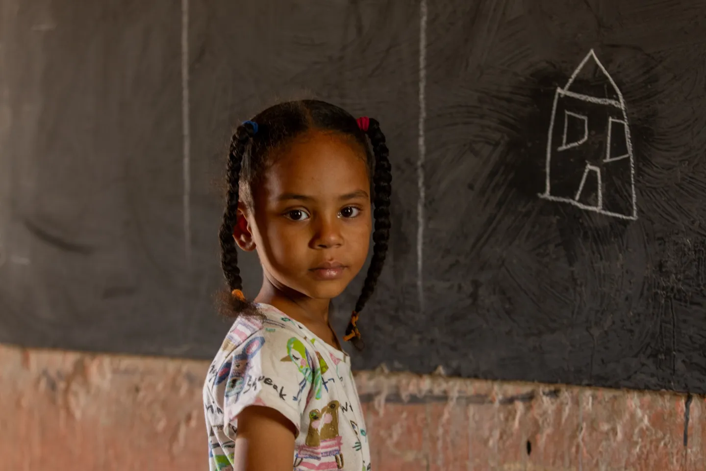 A young girl stands in front of a chalkboard with a picture of a house.