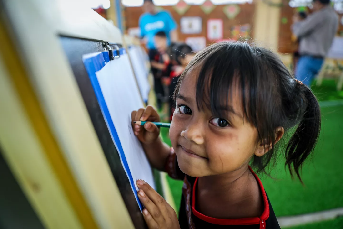 A little girl looks up smiling from where she is drawing on a board.