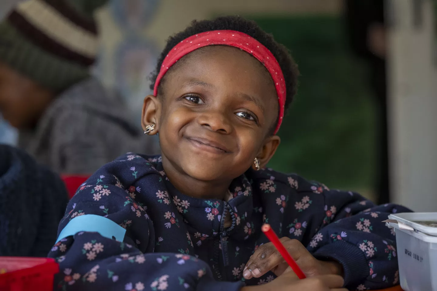 A little girl with a red band in her hair and red pencil in hand smiles happily.
