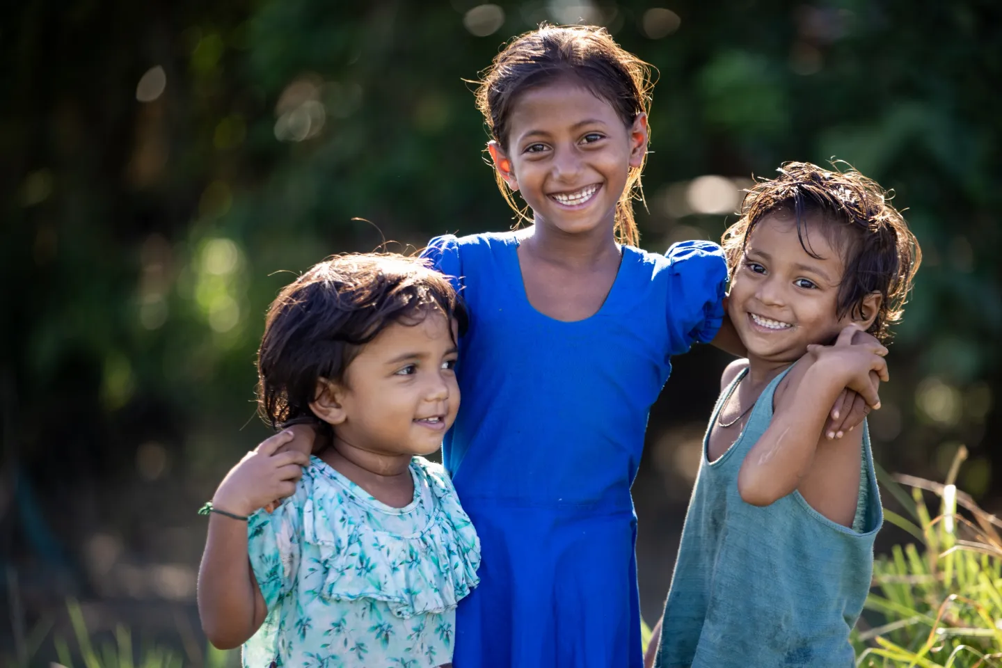Bangladesh: Three little girls smile happily, with the oldest girl in the middle with her arms wrapped around the shoulders of the younger ones.
