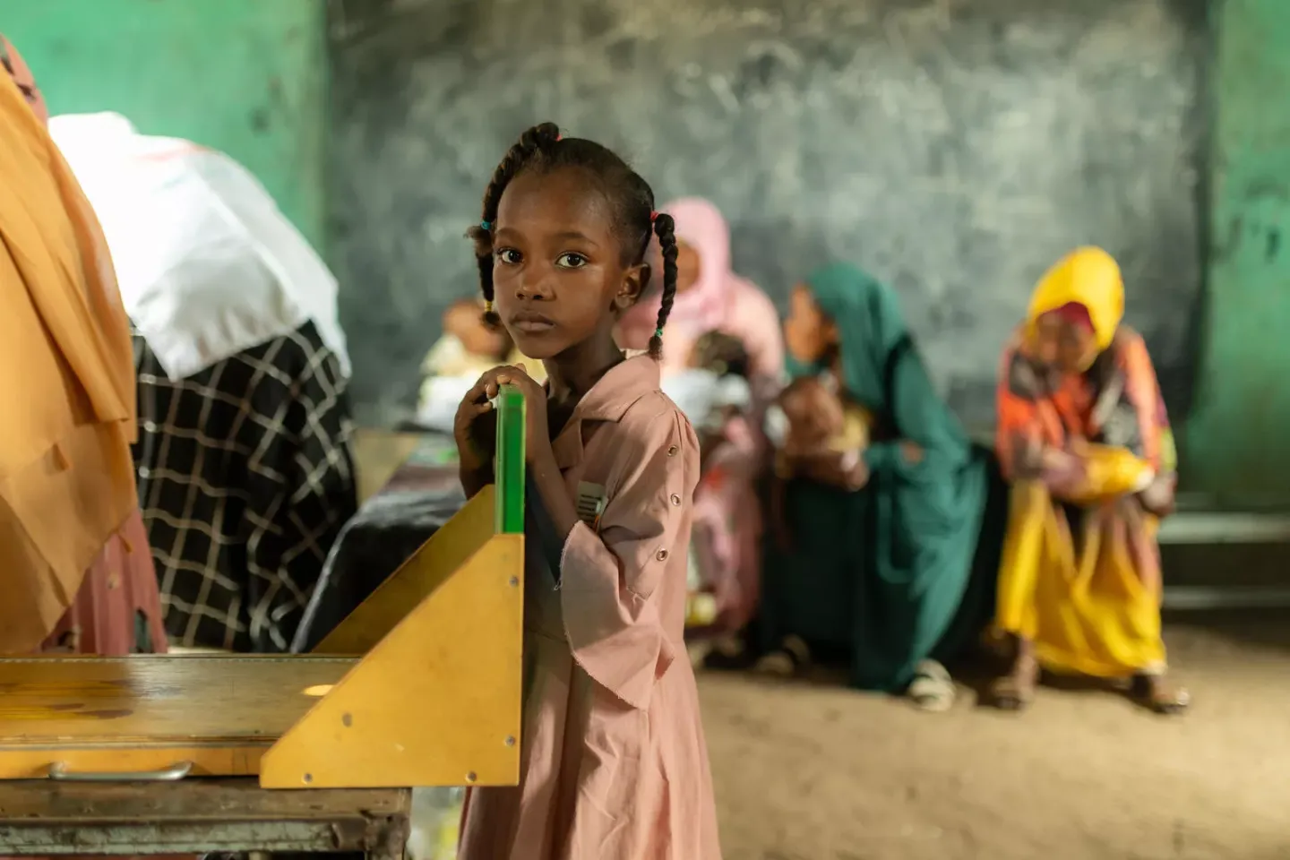 A young girl in a pink dress stands in a classroom in Umshouka Village, Sudan.