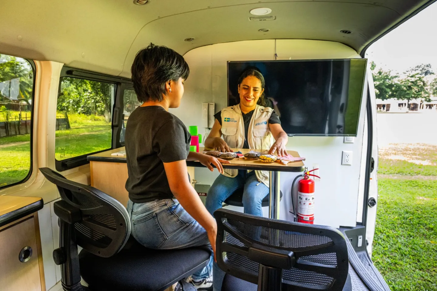 A young woman talks to UNICEF staff member wearing a UNICEF waistcoat, in a mobile meeting space outside.