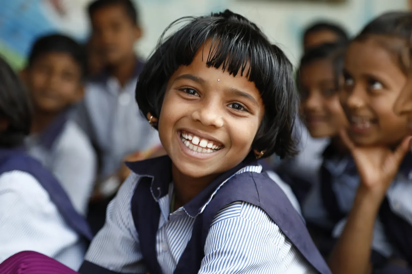 A young girl smiles while sitting in a classroom full of her peers.