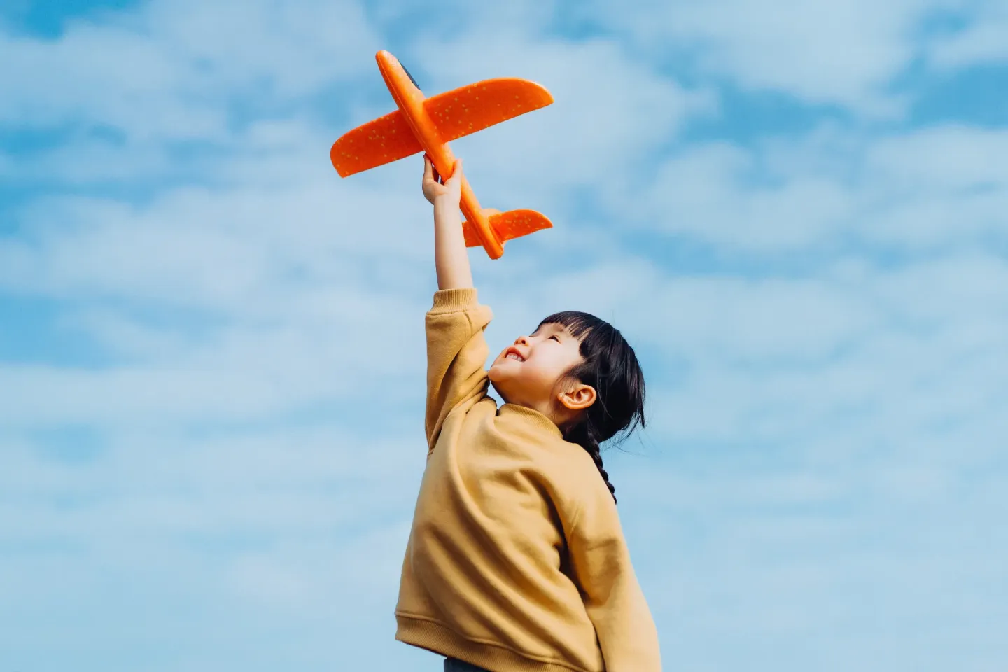 A young girl holds an orange toy airplane high above her head.