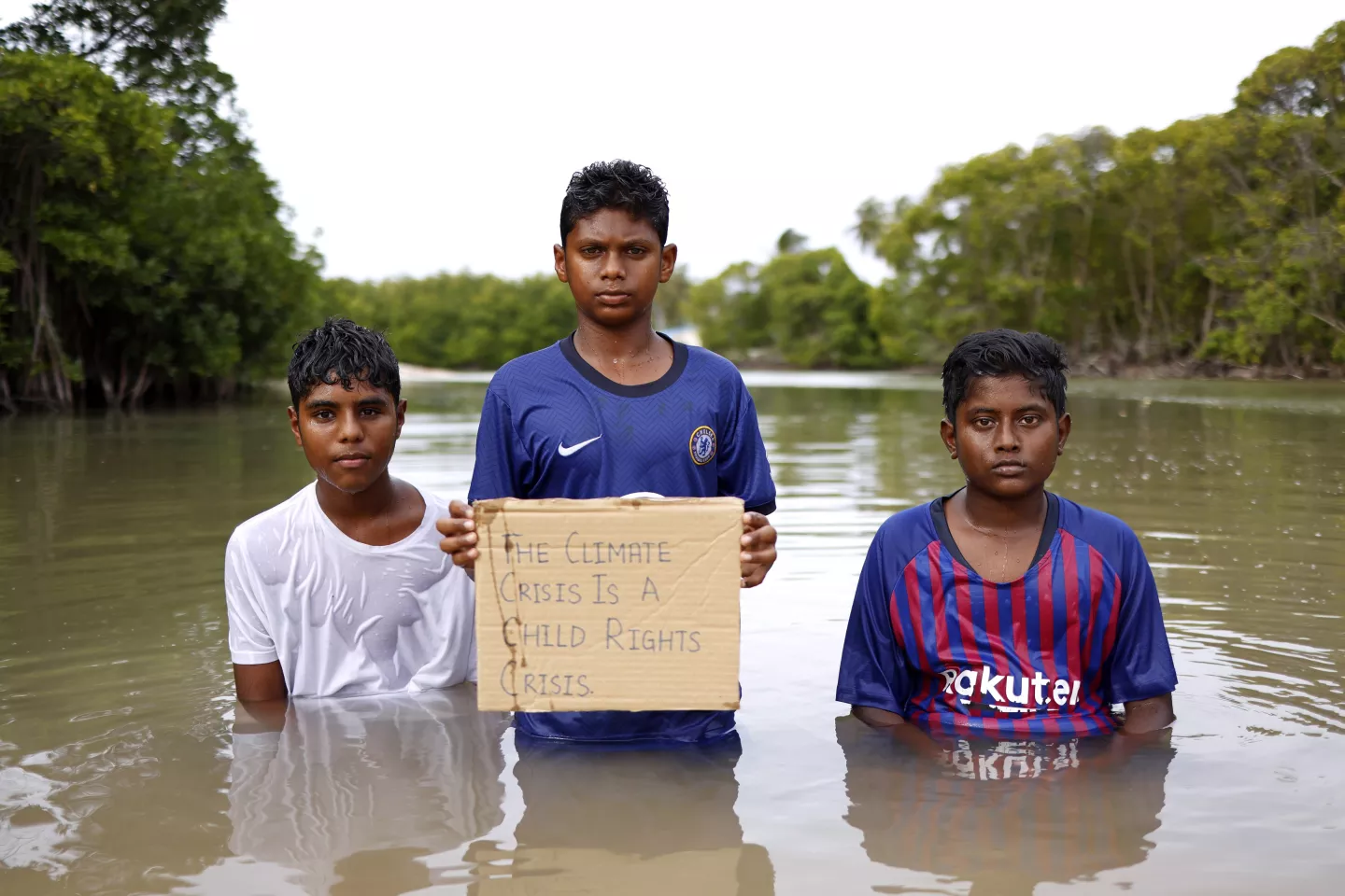 Three boys in flooded plains standing in waist high water. The boy in the middle child has up a placard saying: The Climate Crisis is a Child Rights Crisis