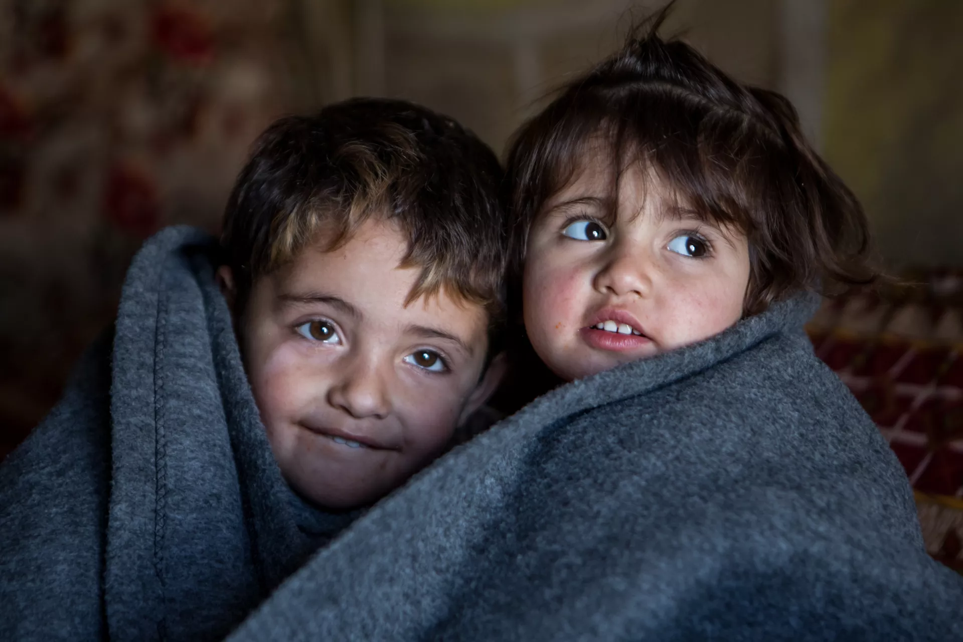 Two children nestle beside each other, wrapped in a thick grey blanket inside their family's tent in a refugee camp in Iraq.