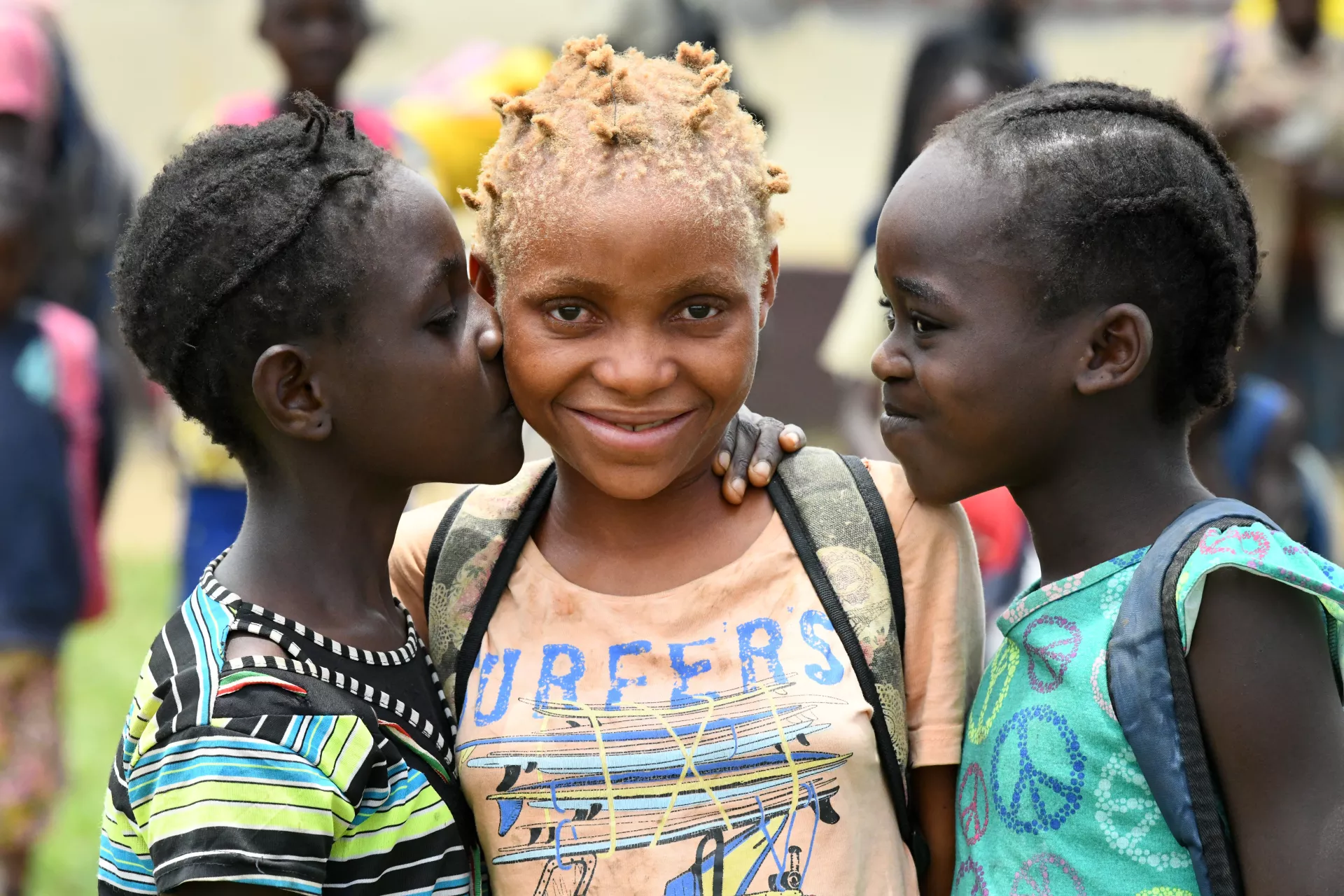 3 jeunes filles sourient. L'une embrasse l'autre sur la joue.