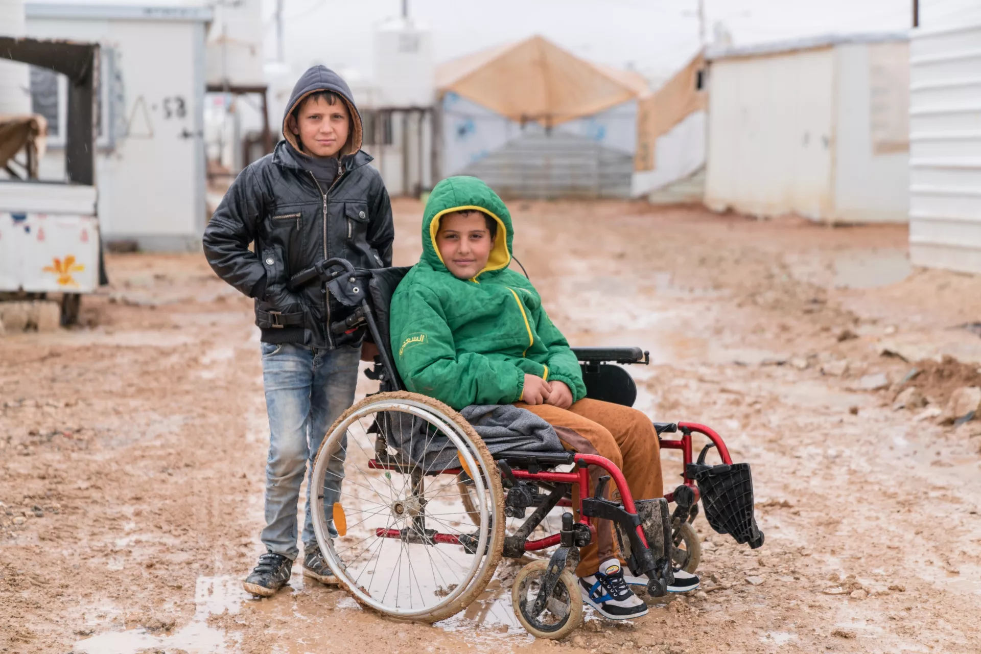 Two boys, one standing, one in a wheelchair smile for the camera in refugee camp