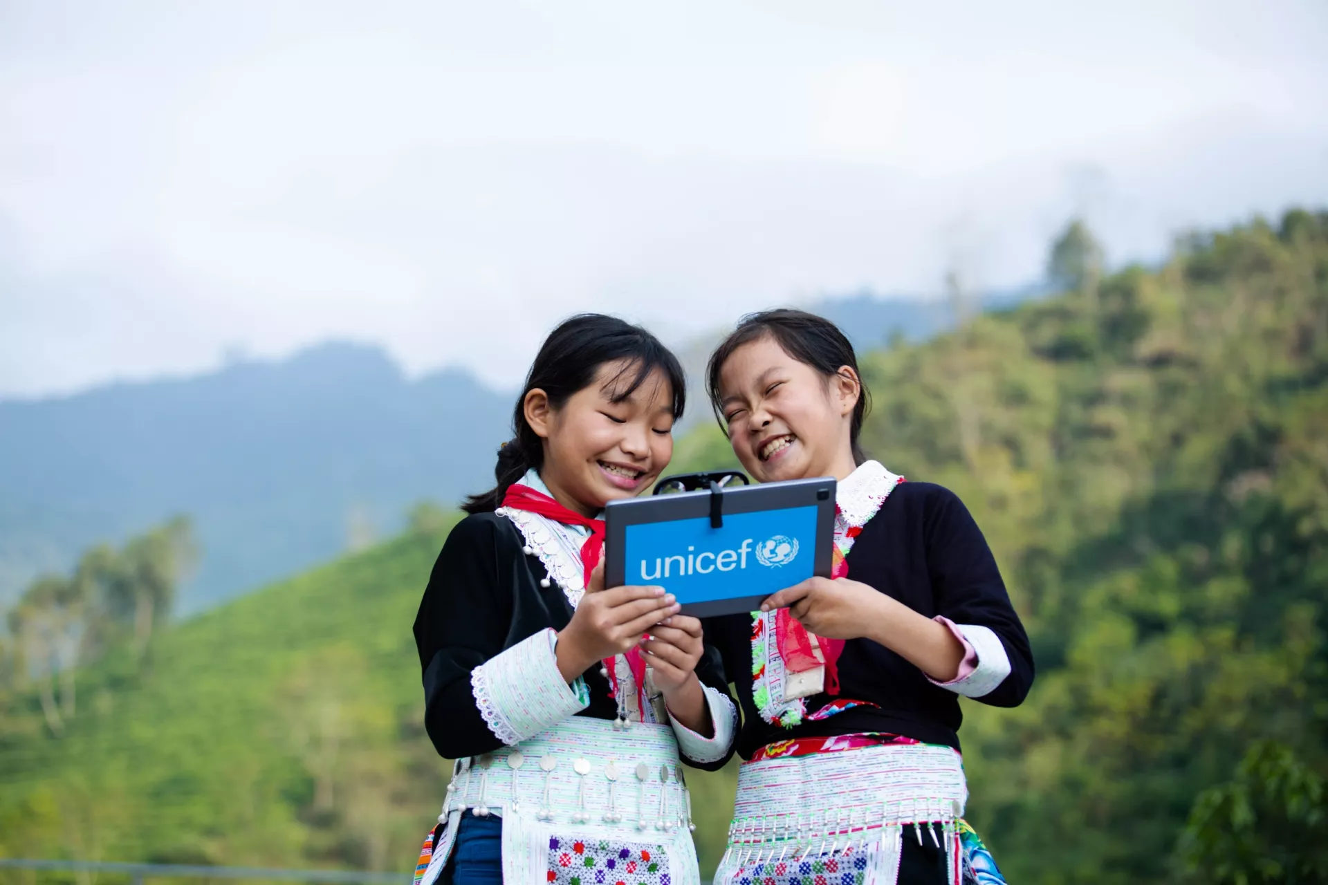 Two girls dress in traditional clothing laugh over something that they see on the tablet that they are holding.