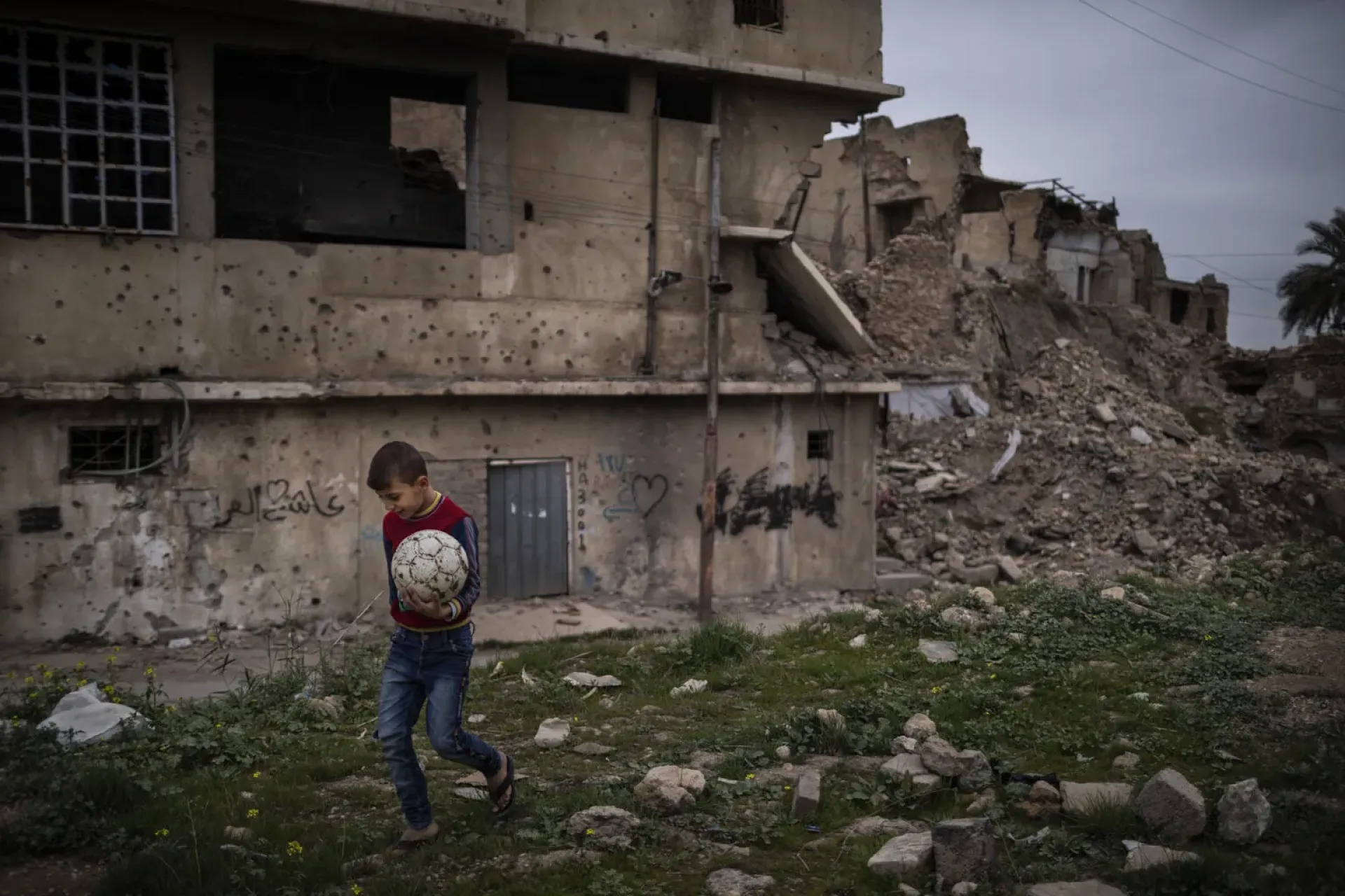 Iraq. A boy holding a football in one hand walks past a damaged building.