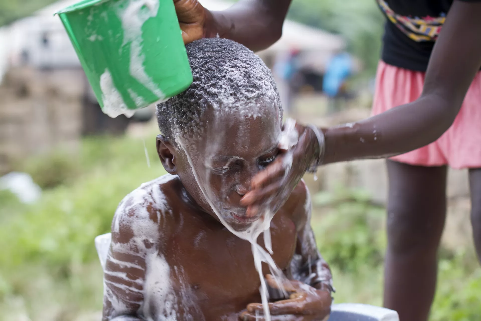 A boy covered in soap sits in a tub, while his sister pours water over him from a bucket and washes his face with her other hand.