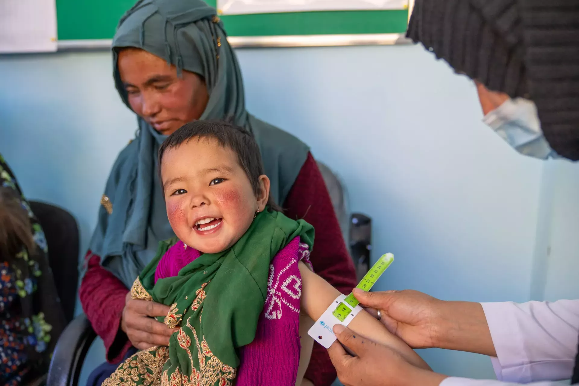 A girl sitting on her mom's lap smiles broadly while her mid-upper arm circumference is measused.