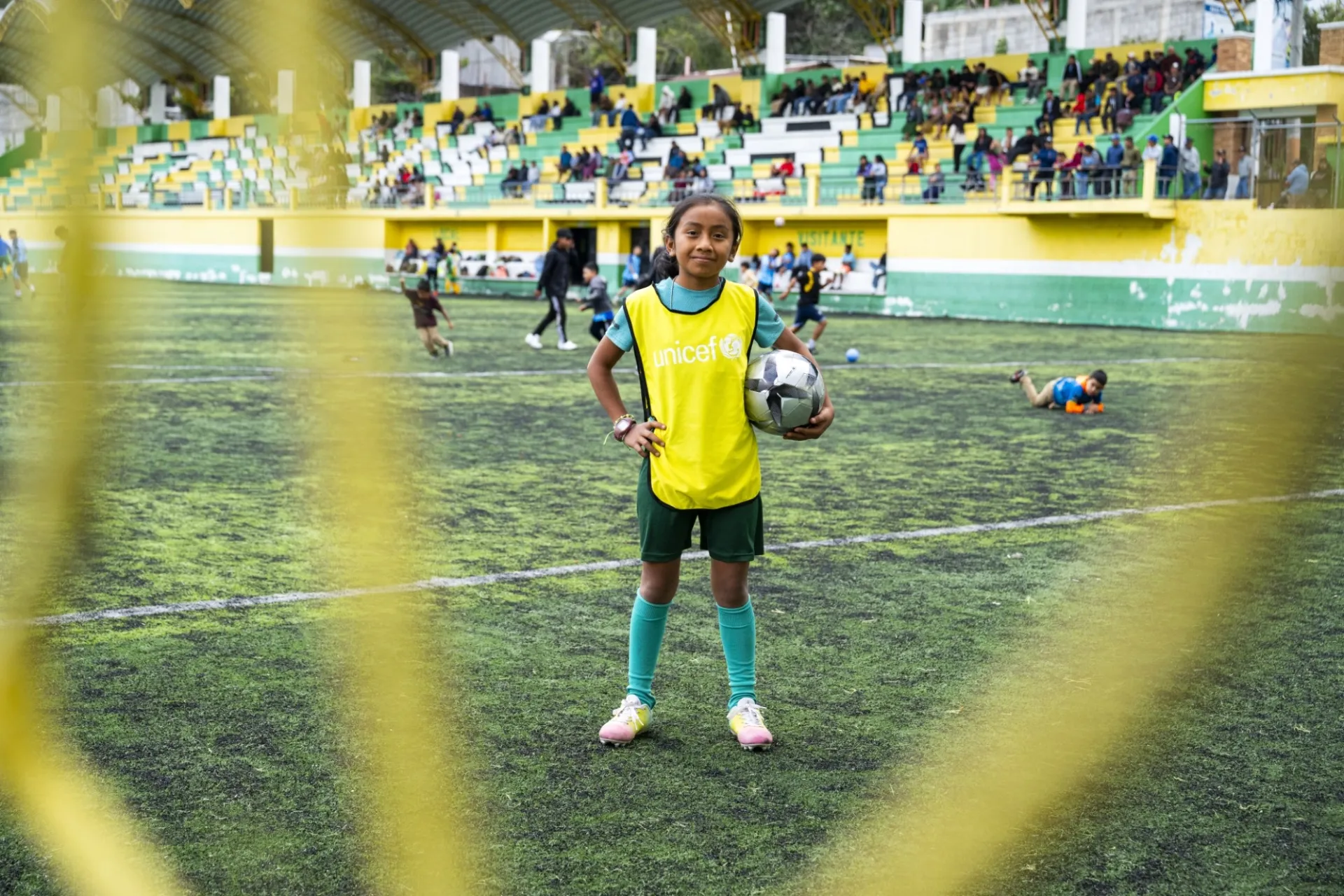 Guatemala: A girl stands on a football pitch, holding a ball under her arm. She is wearing football clothes with a yellow UNICEF team bib over it.
