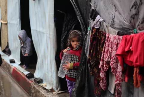 On a rainy day, a young girl stands in front of her shelter in Rafah while holding an empty water bottle.