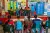 A group of children sit in a row on a stage with a UNICEF banner to the side, with a row of children looking on.