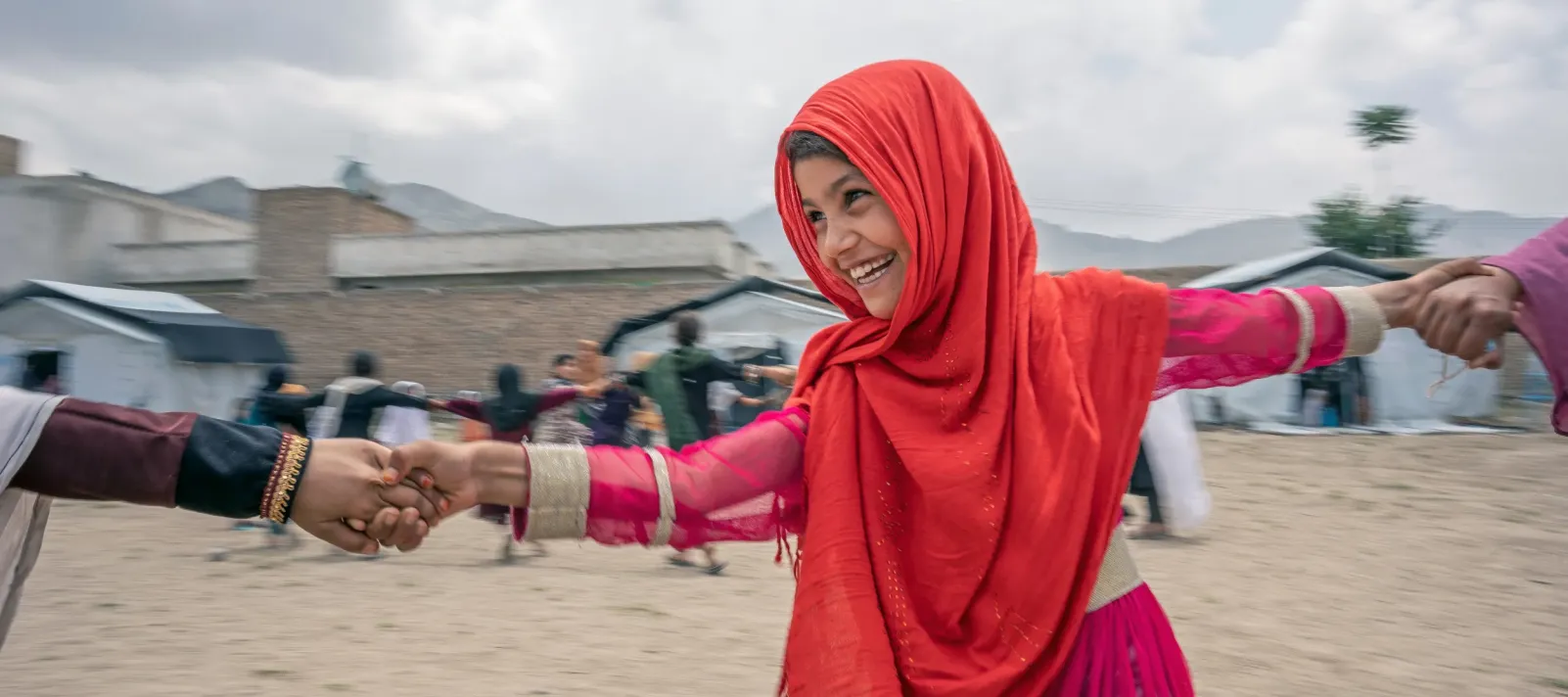 A girl in a red scarf smiles brightly while looking towards a friend, holding hands while playing.