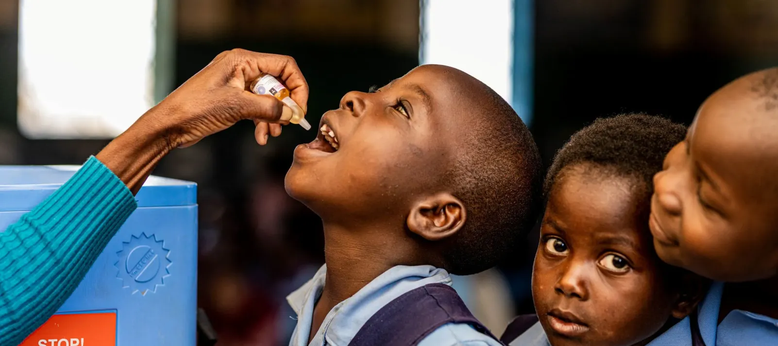 A child receives an oral poli vaccine while two young peers look on.