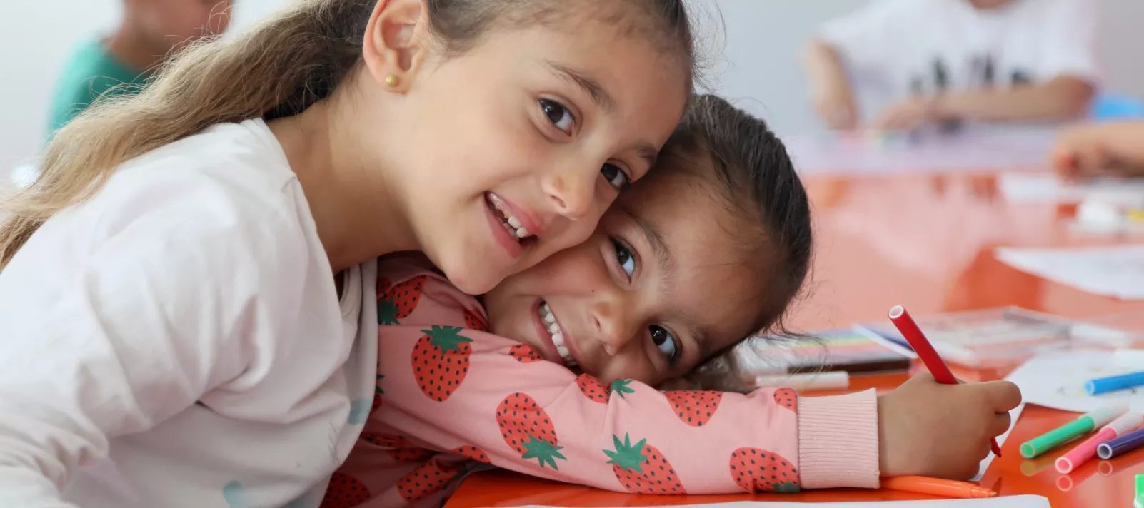 A young girl leans over her friend smiling while sitting at a table drawing.