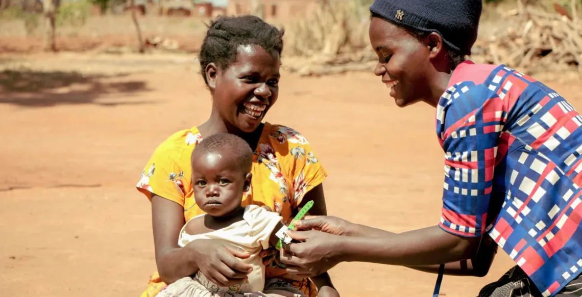 A woman holds a child on her lap while another woman measures the upper arm of the child