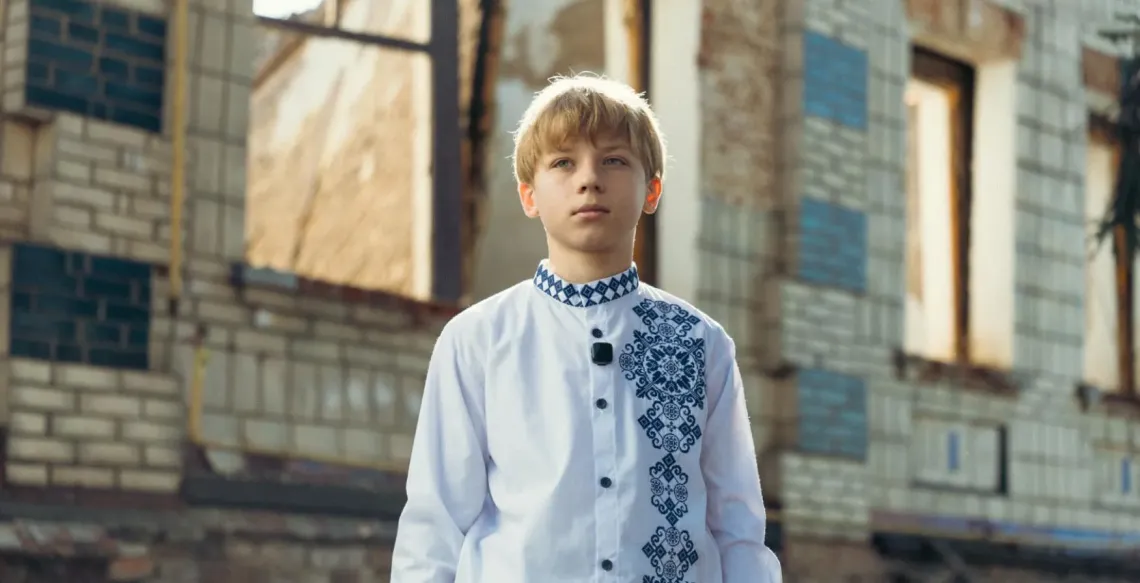 An adolescent boy stands in front of a damanged building