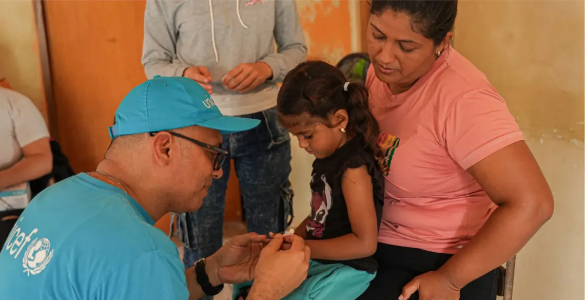 A girl sits on a woman&rsquo;s lap and a man in UNICEF-branded clothing is in front of them