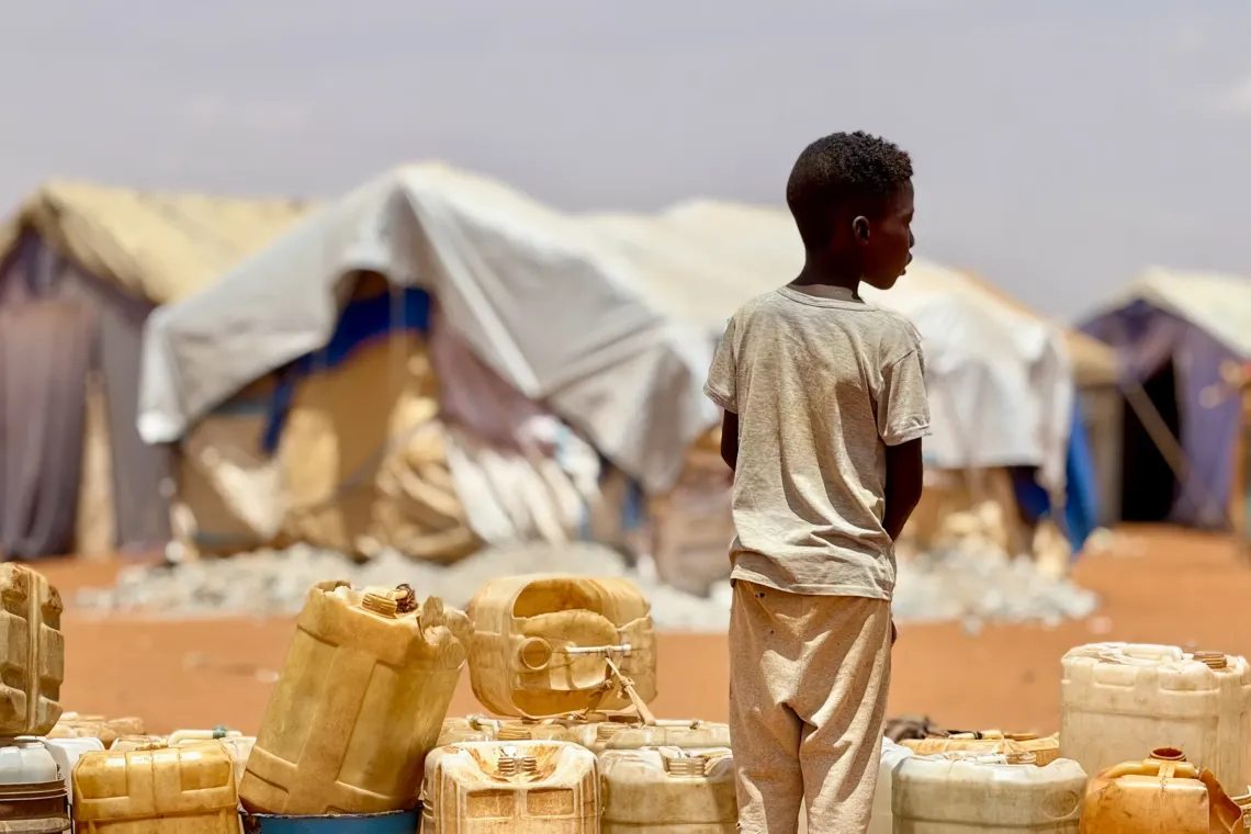 A boy stands alongside empty water jugs, with shelter tents in the background.