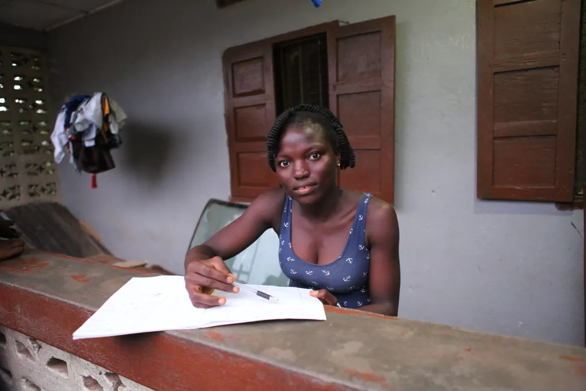 A teenage girl sits at a desk and writes in a notebook, Sierra Leone