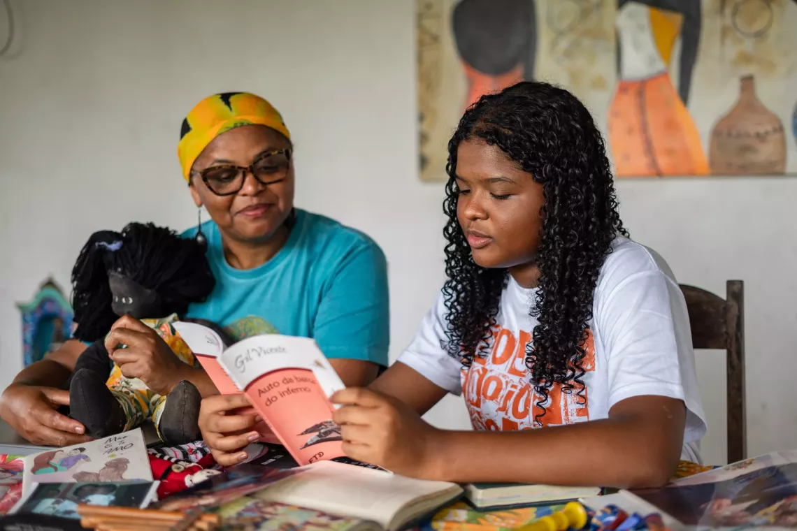 A girl and her mother sit next to each other at a table. The girl reads from a book, while her mother listens attentively.